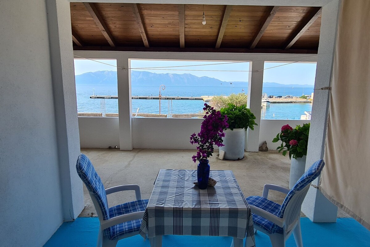 A covered patio with a table and chairs, a vase with flowers, and a view of the sea and mountains.