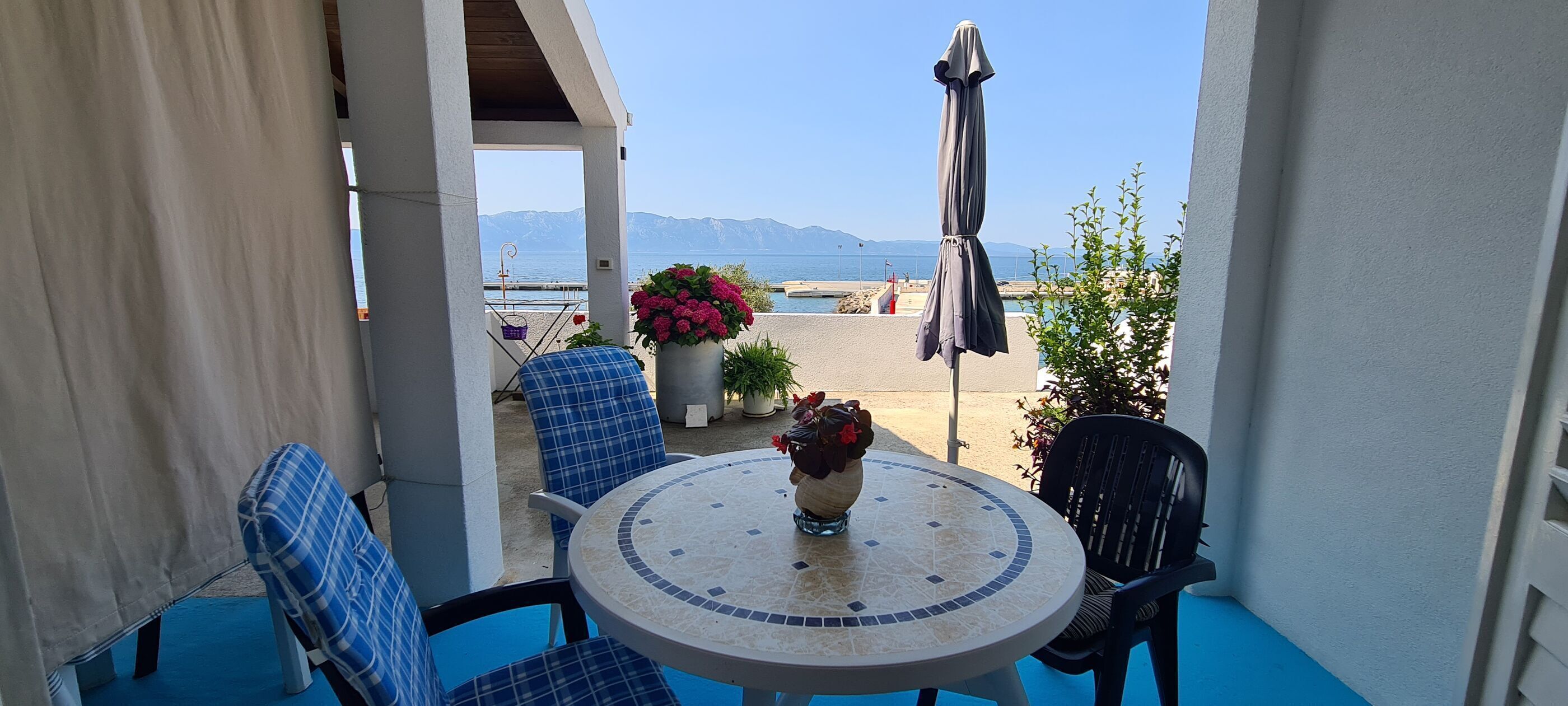 Un patio con un tavolo rotondo, sedie blu, una pianta in vaso e vista sul mare e sulle montagne.