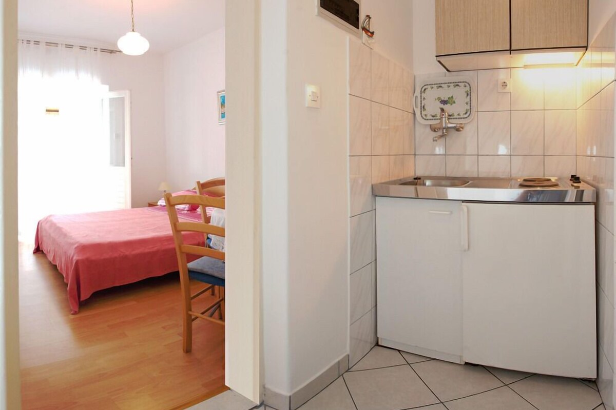A compact bedroom with a bed and a chair, a small kitchenette with a sink and cabinets, and a door leading to another room.