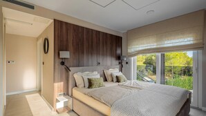 Ein Schlafzimmer mit einem großen Bett, einer Wandverkleidung aus Holz, einem Nachttisch, einer Wandlampe und Blick auf Grün hinter einer Schiebetür.