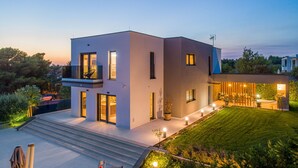 Ein modernes, zweistöckiges Haus mit Flachdach, großen Fenstern und einem gut beleuchteten Außenbereich mit Rasen und Sitzgelegenheit.