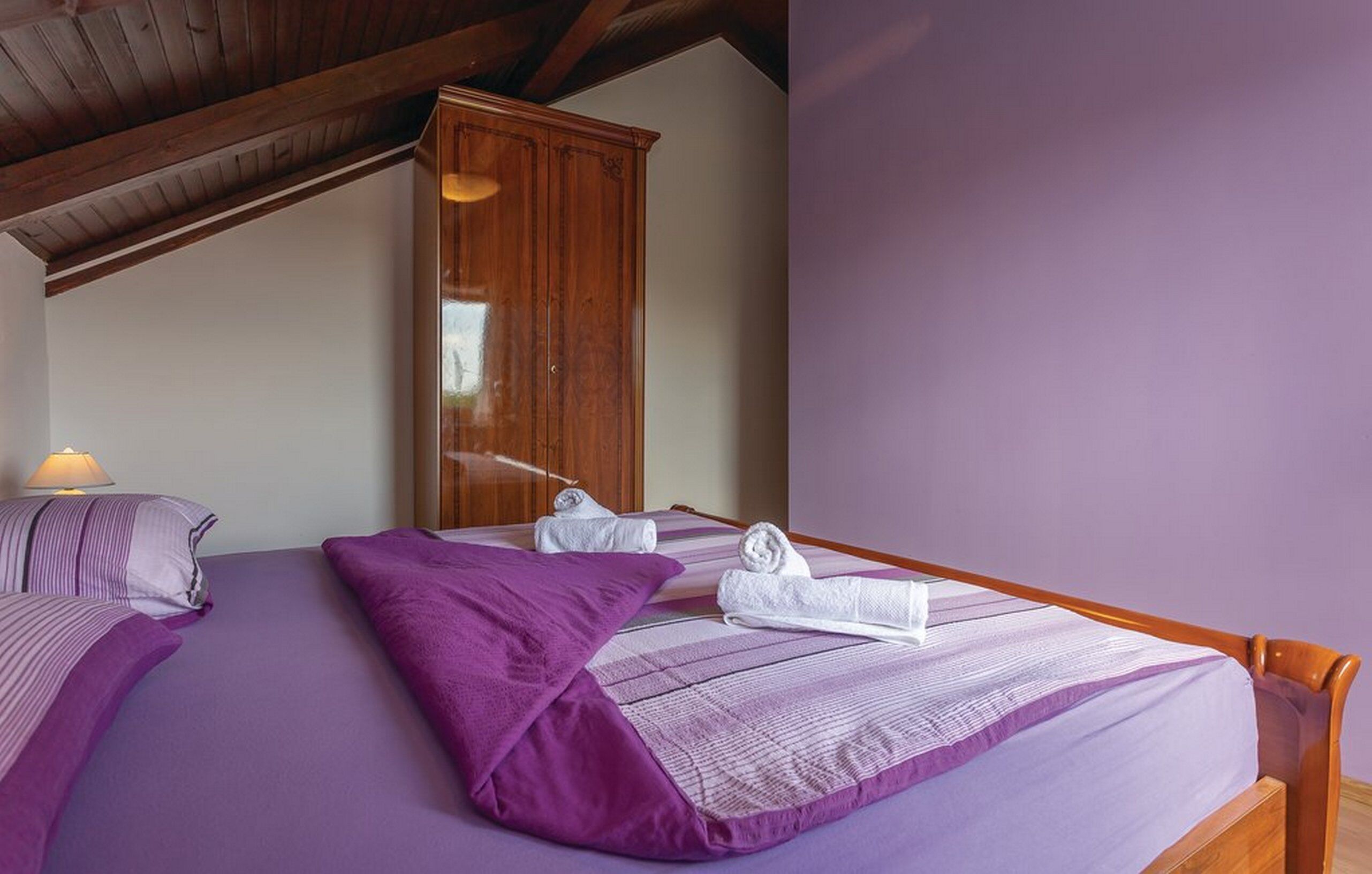 A bedroom with two beds, purple bedding, wooden headboards, a wardrobe, and a lamp.