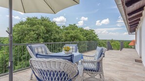 A balcony with wicker furniture, a table with a vase of flowers, and a view of greenery and a distant building. - House Rural Soul (Promina)