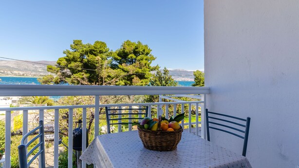 Ένα μπαλκόνι με ένα τραπέζι και καρέκλες, ένα καλάθι από μπαμπού γεμάτο με φρούτα και θέα στη θάλασσα και στα δέντρα.