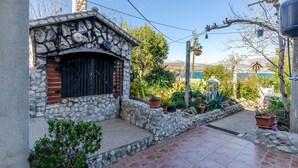 Ένα πέτρινο σπίτι με μια μαύρη πύλη, περιτριγυρισμένο από φυτά σε γλάστρες και μια πλακόστρωτη αυλή.