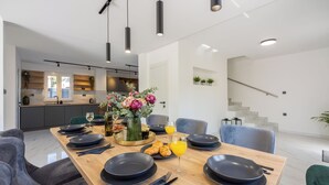 A modern dining area with a wooden table set for a meal, a kitchen in the background, and a staircase leading to another level. - House Kety (Baska)