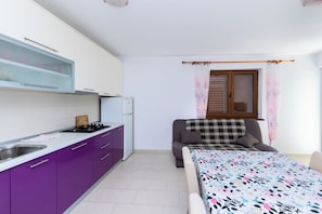 Una habitación compacta con kitchenette, una cama con colcha estampada y una ventana con cortinas.