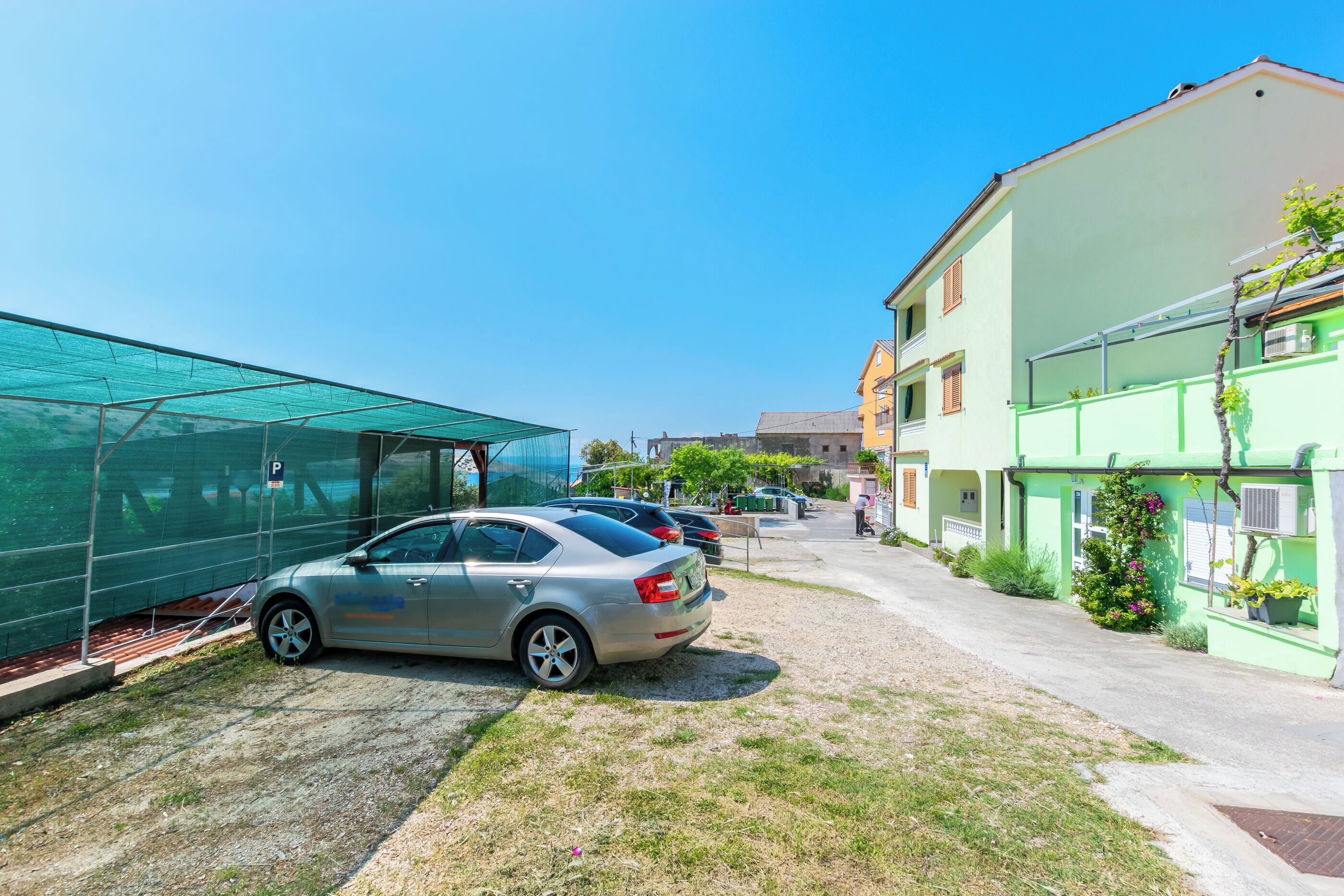 A parking area with a car, a green structure, and a building with a balcony.