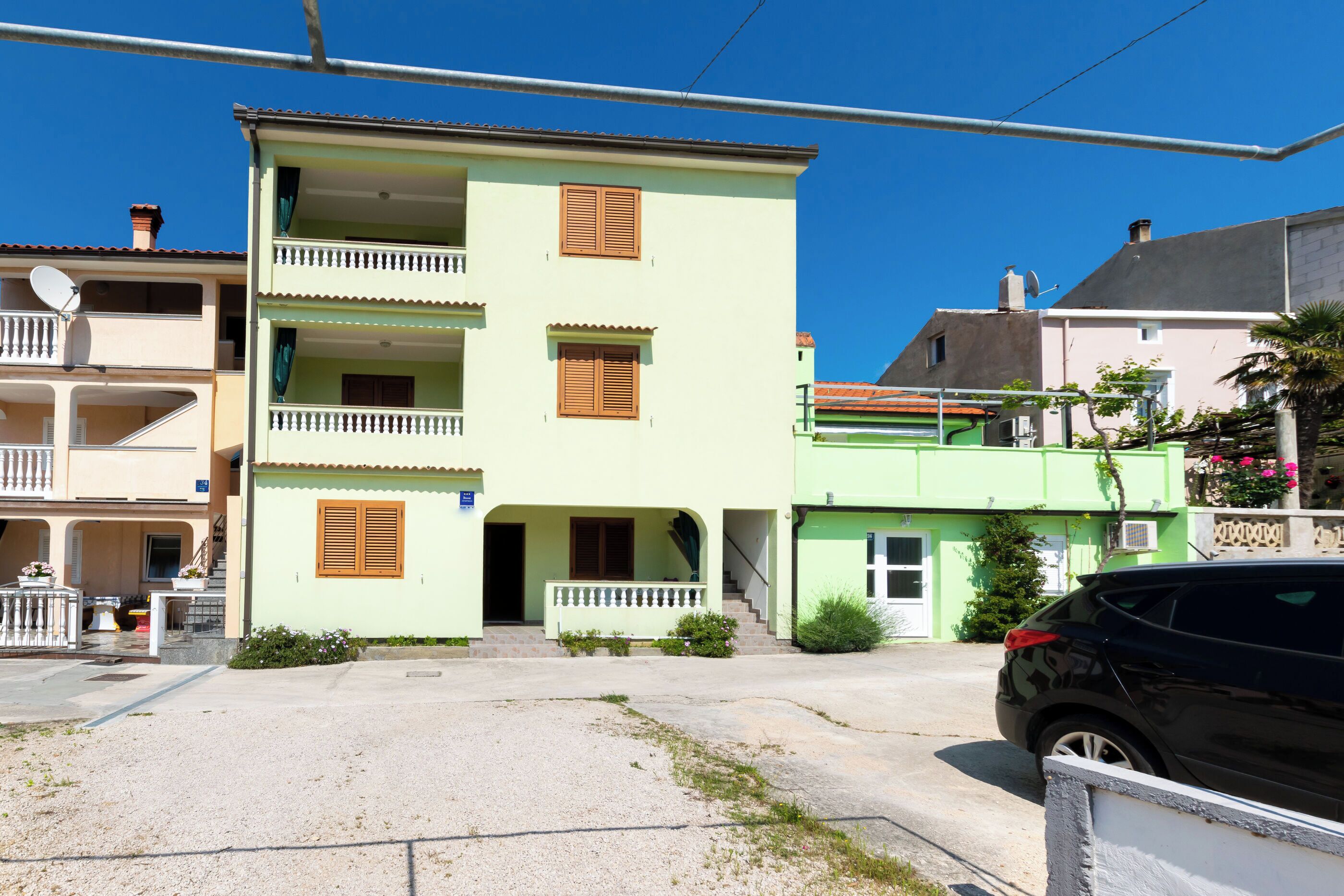 A multi-story building with balconies, a car parked in front, and a clear blue sky.