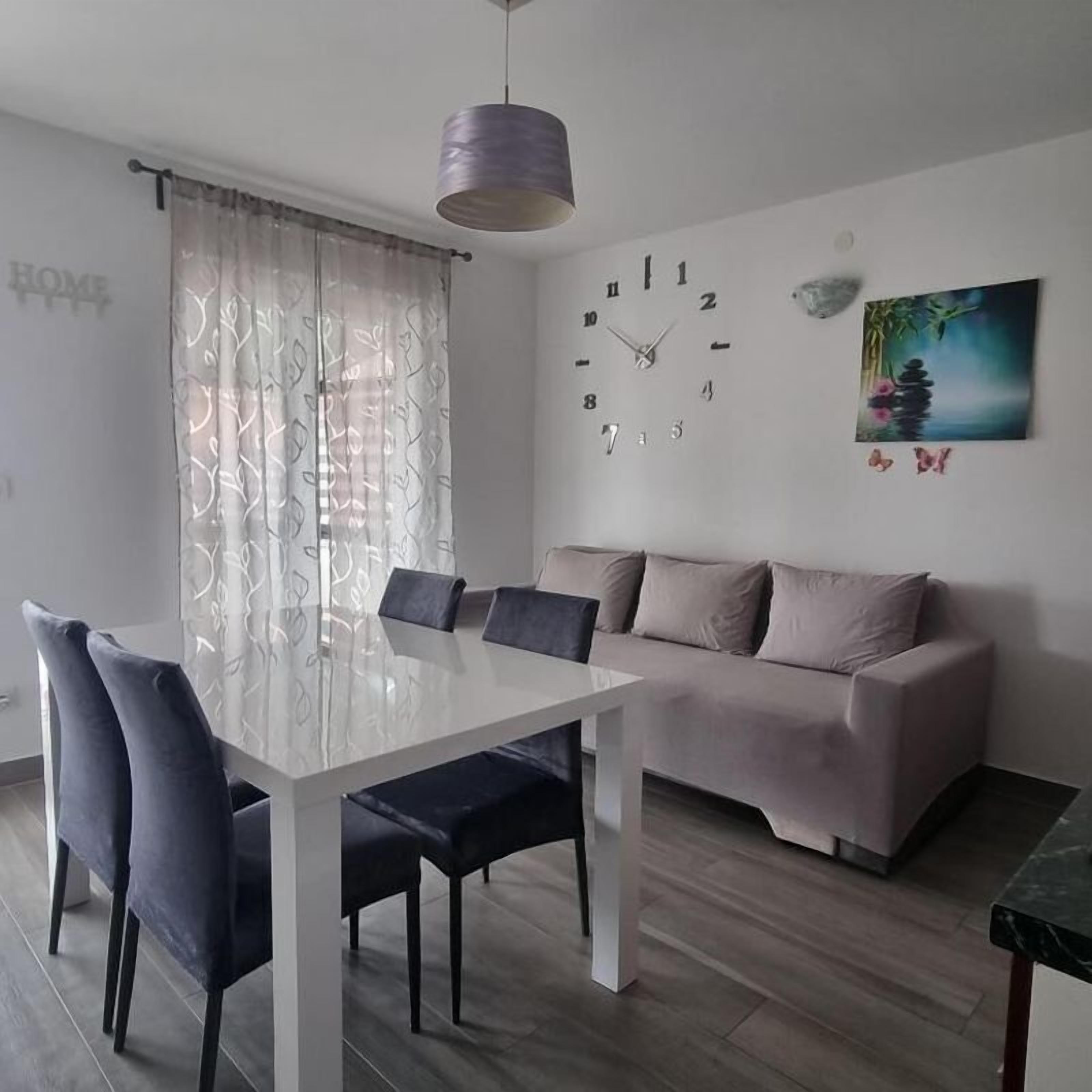 A modern living room with a white sofa, a dining table with chairs, a wall clock, and a painting.
