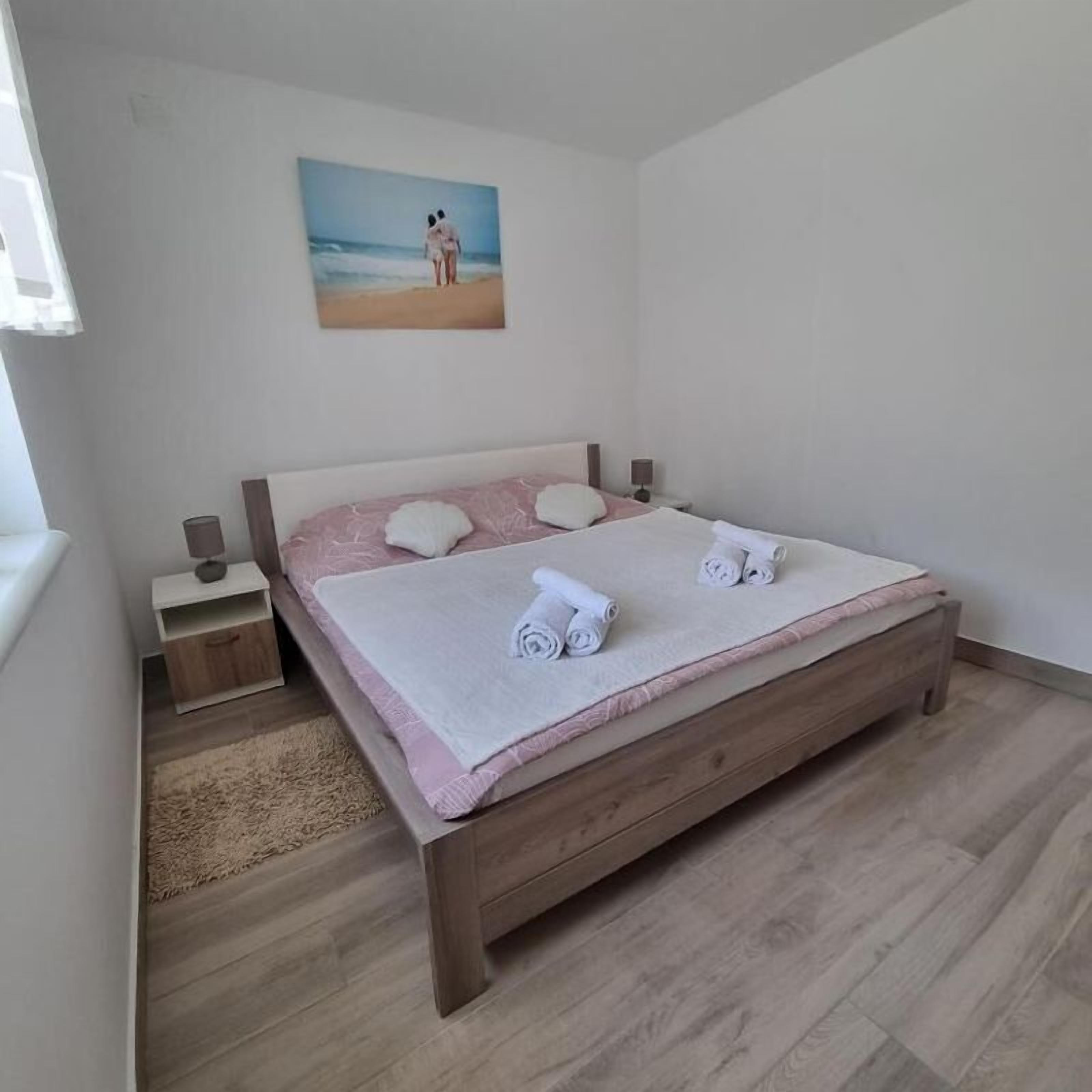 A bedroom with a bed, a nightstand, a lamp, a painting of a beach scene, and a wooden floor.