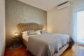 Un dormitorio con cabecera capitonada, cama con colcha gris, lámpara de noche y alfombra redonda.