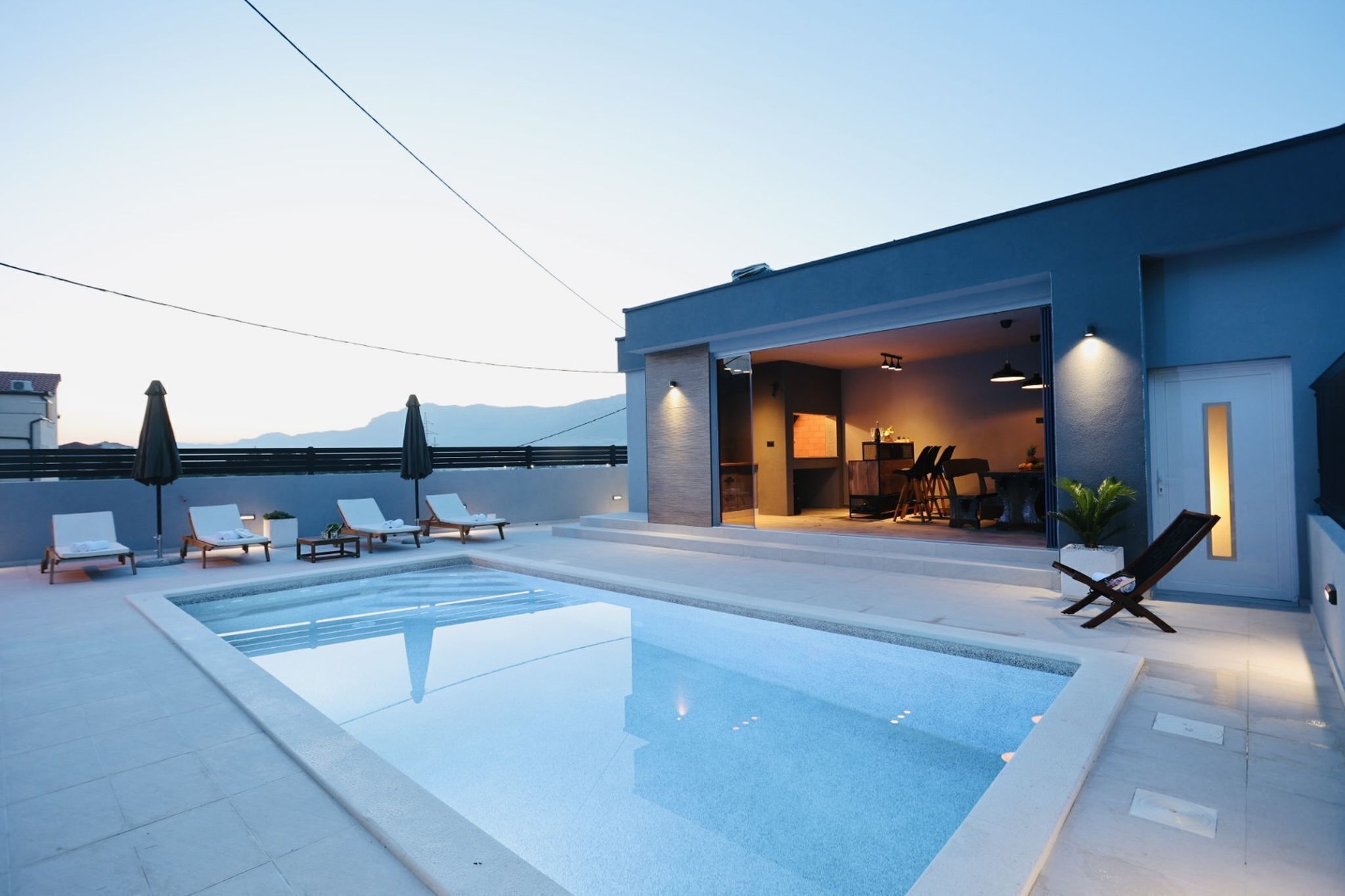 Zona de piscina moderna amb gandules i ombrelles, interior ben il·luminat i vistes a les muntanyes de fons.