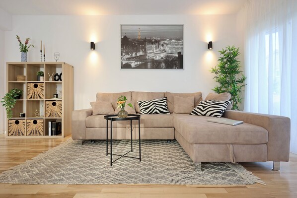 Una sala de estar moderna con un sofá color beige, una mesa de centro negra y un tapete estampado. Se observa una estantería con adornos y una fotografía en grande de un paisaje urbano.