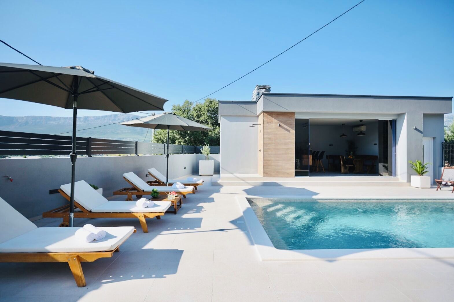 A rooftop pool area with lounge chairs and umbrellas, a modern building with an open entrance, and a clear view of the mountains.