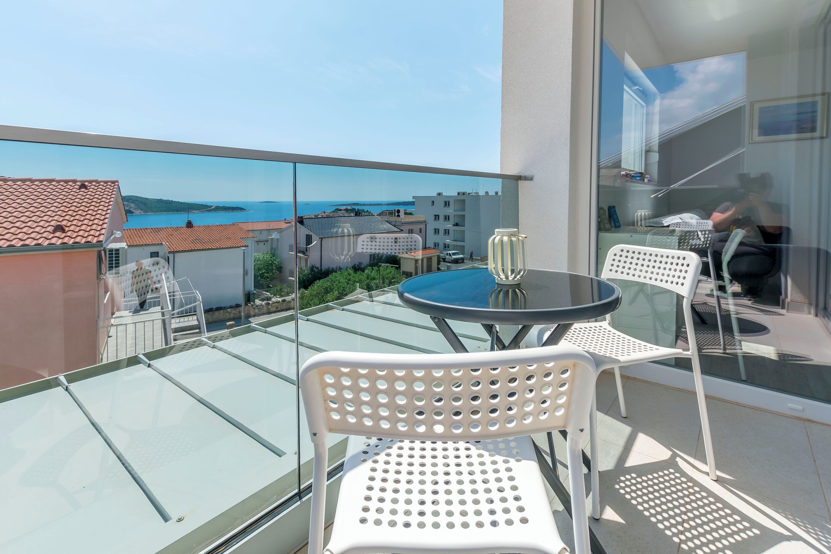Ein Balkon mit Glasboden, weißen Stühlen, einem runden Tisch und Blick auf das Meer und die Gebäude.