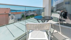 A balcony with a glass floor, white chairs, a round table, and a view of the sea and buildings. - Apartments Tonka (Primosten)