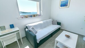 A modern, minimalist living room with a white desk, a green sofa, a small white table, and a window with a view of the sea. - Apartments Tonka (Primosten)