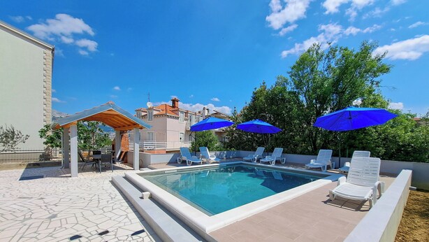A swimming pool with lounge chairs and blue umbrellas, surrounded by a tiled patio and a building with a tiled roof. - Apartments Mile (Split)