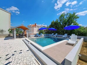 A swimming pool with lounge chairs and blue umbrellas, surrounded by a tiled patio and a building with a tiled roof. - Apartments Mile (Split)