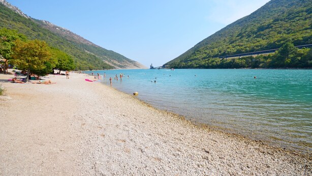 A pebble beach with a clear lake, a bridge, and hills in the background. - Apartments Plomin (Krsan)