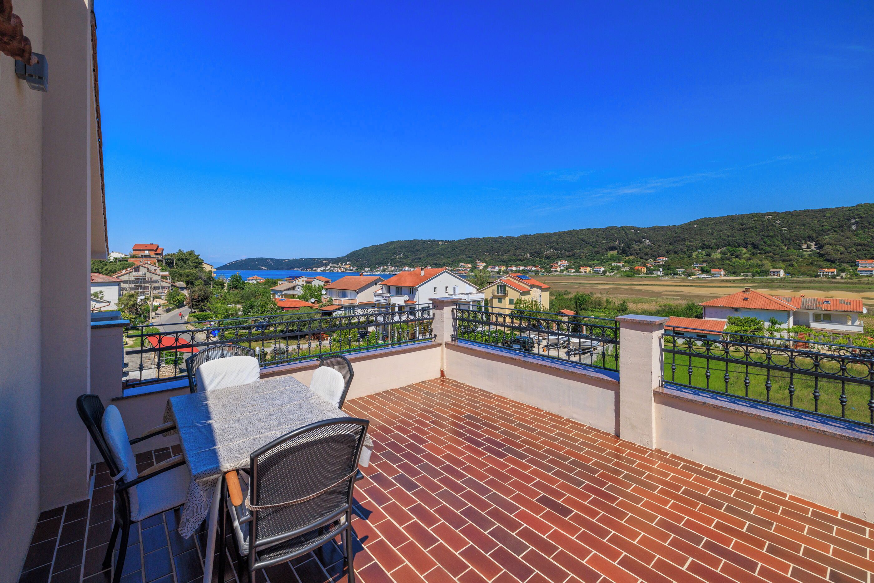 A balcony with a table and chairs, overlooking a residential area with houses and a clear blue sky.