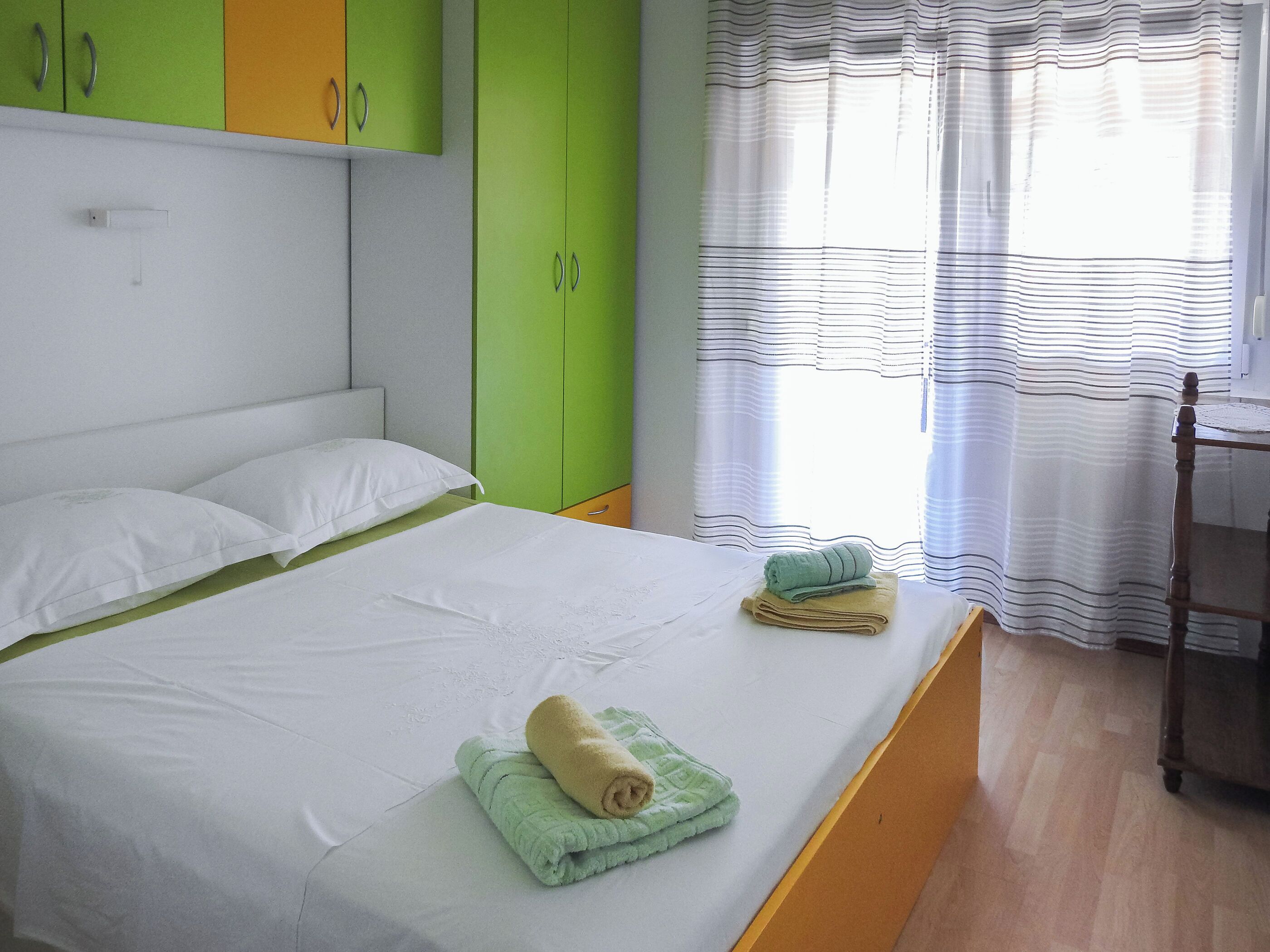 A bedroom with a bed, green and yellow cabinets, a wooden chair, and a window with striped curtains.