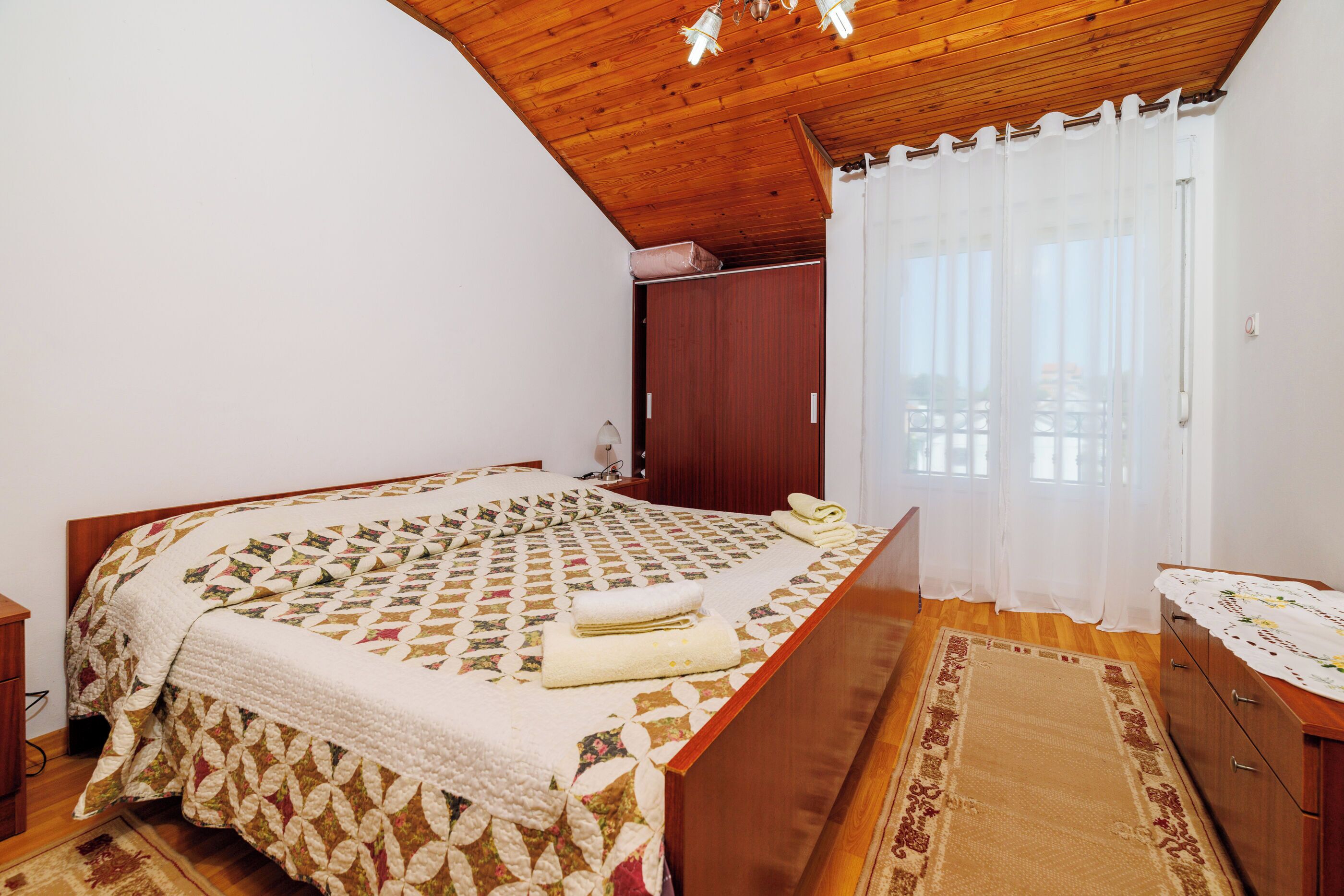 A bedroom with a wooden ceiling, a bed with a patterned quilt, a wooden dresser, and a window with white curtains.