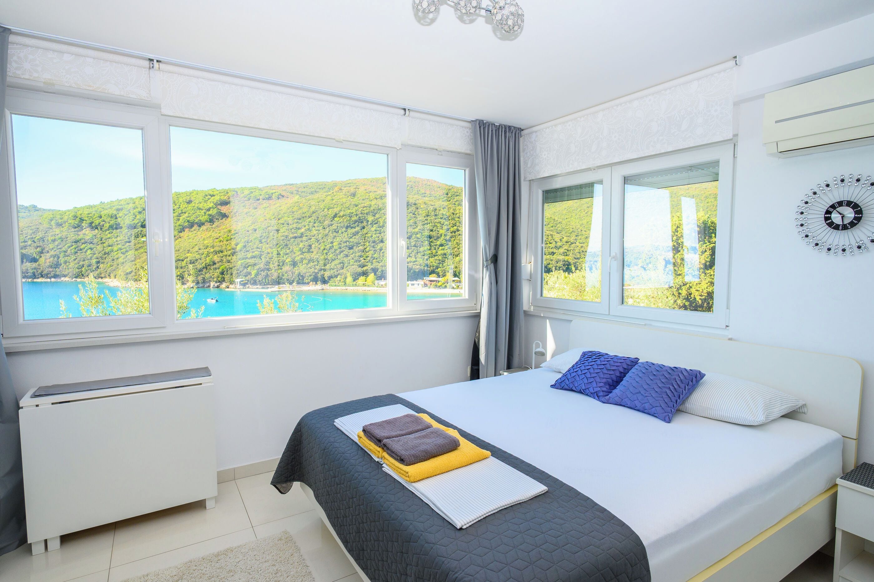 Un dormitorio con cama, mesita de noche, un reloj de pared y vistas a un lago y colinas.