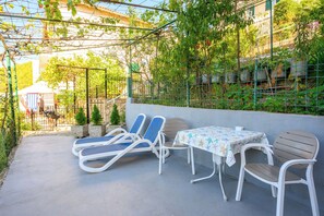 An outdoor seating area with lounge chairs and a table, surrounded by greenery and a metal fence. - Apartment By the Sea (Labin)