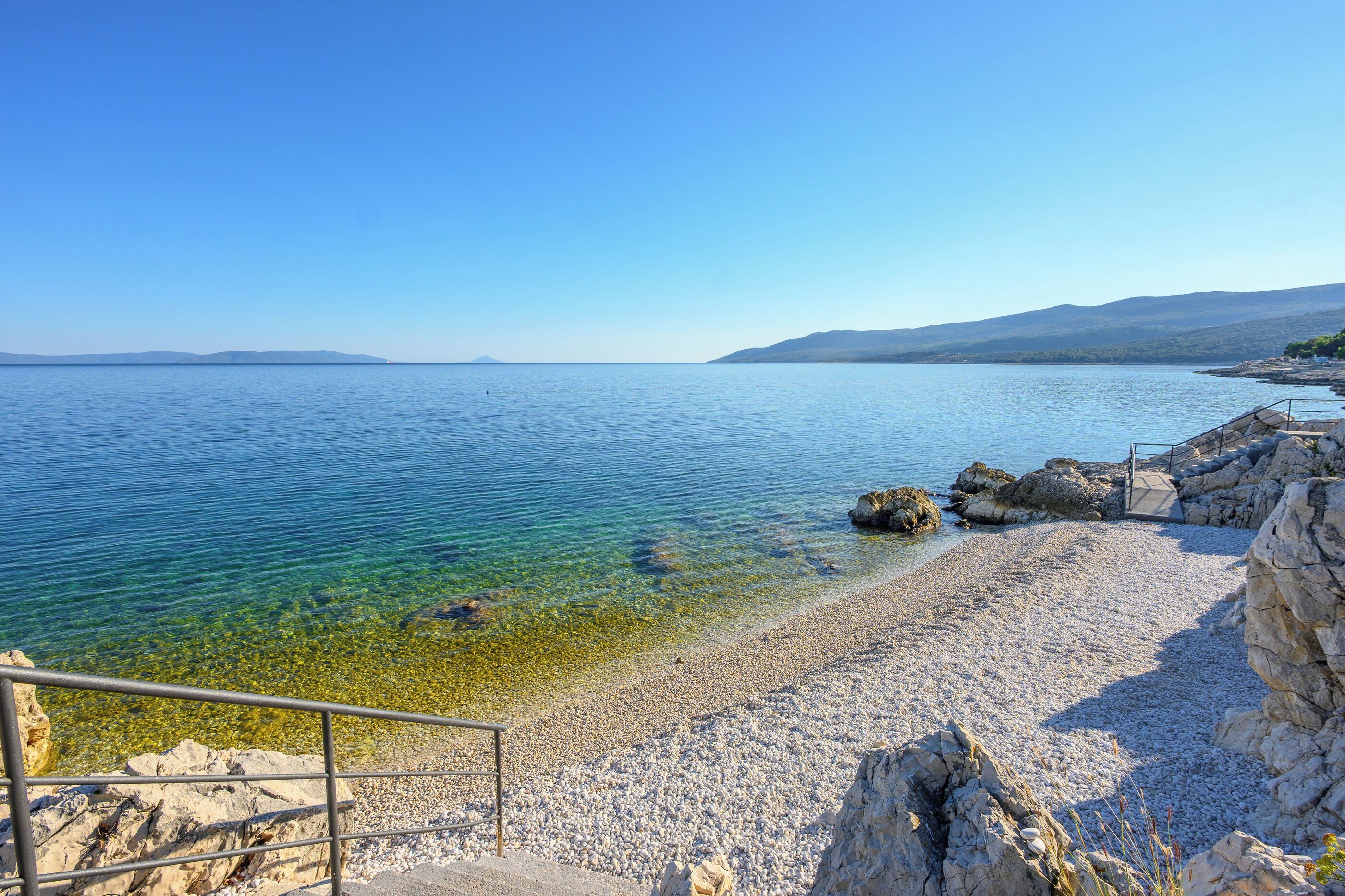 Une plage de galets bordée d’une eau cristalline, un promontoire rocheux et, au loin, une chaîne de montagnes.