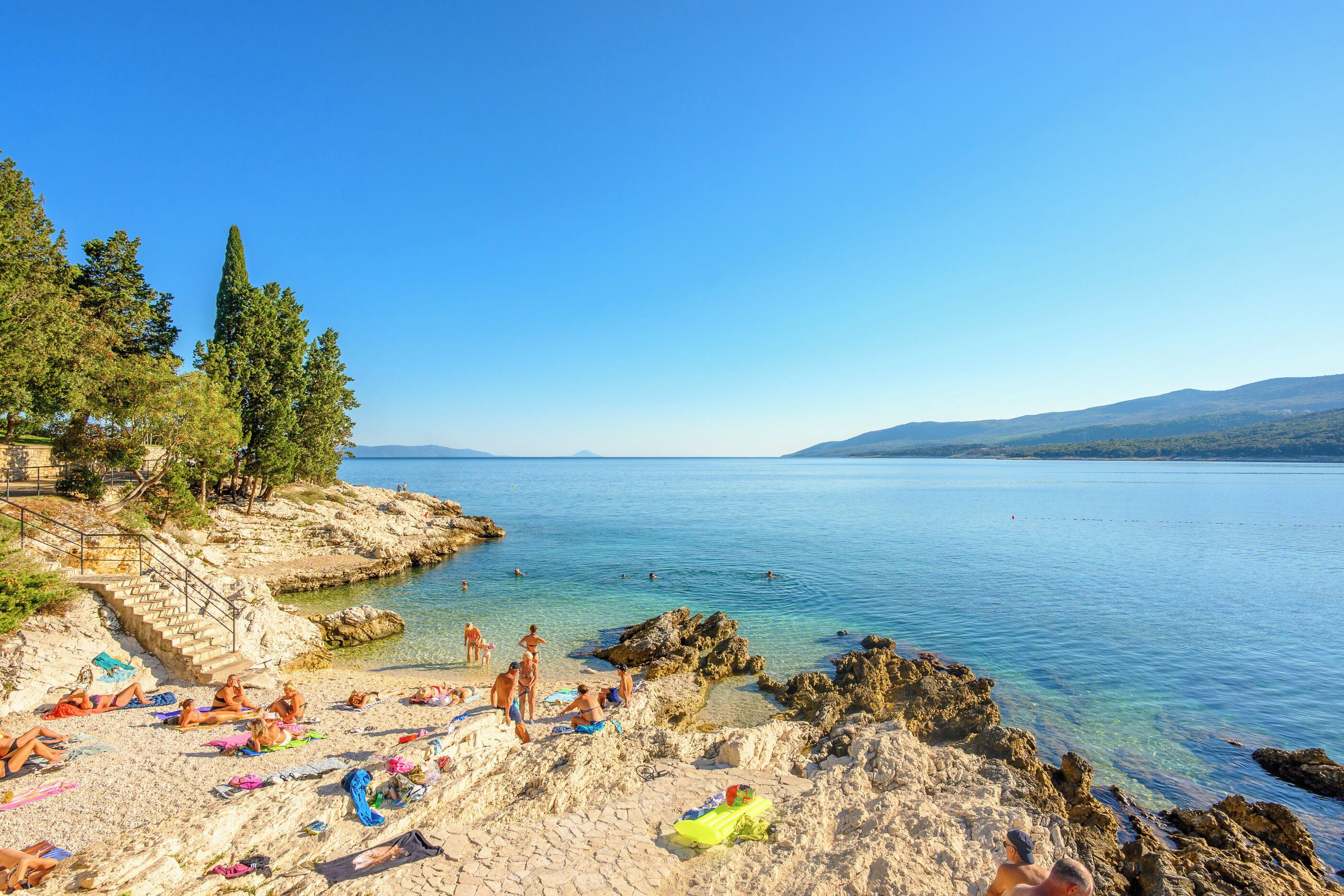 A coastal beach with clear water, rocky terrain, and trees.