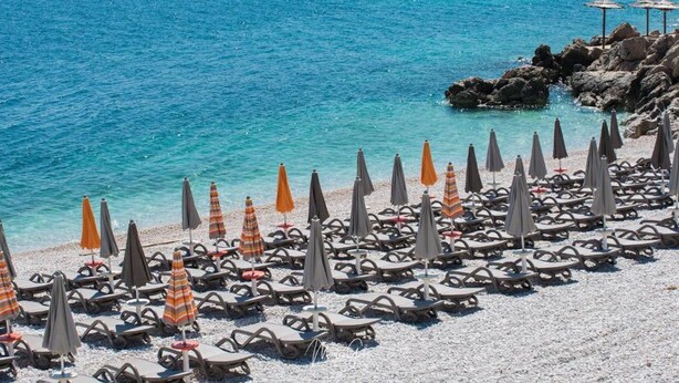 Een strand met rijen ligbedden en parasols, een rotsachtige kustlijn en helderblauw water.
