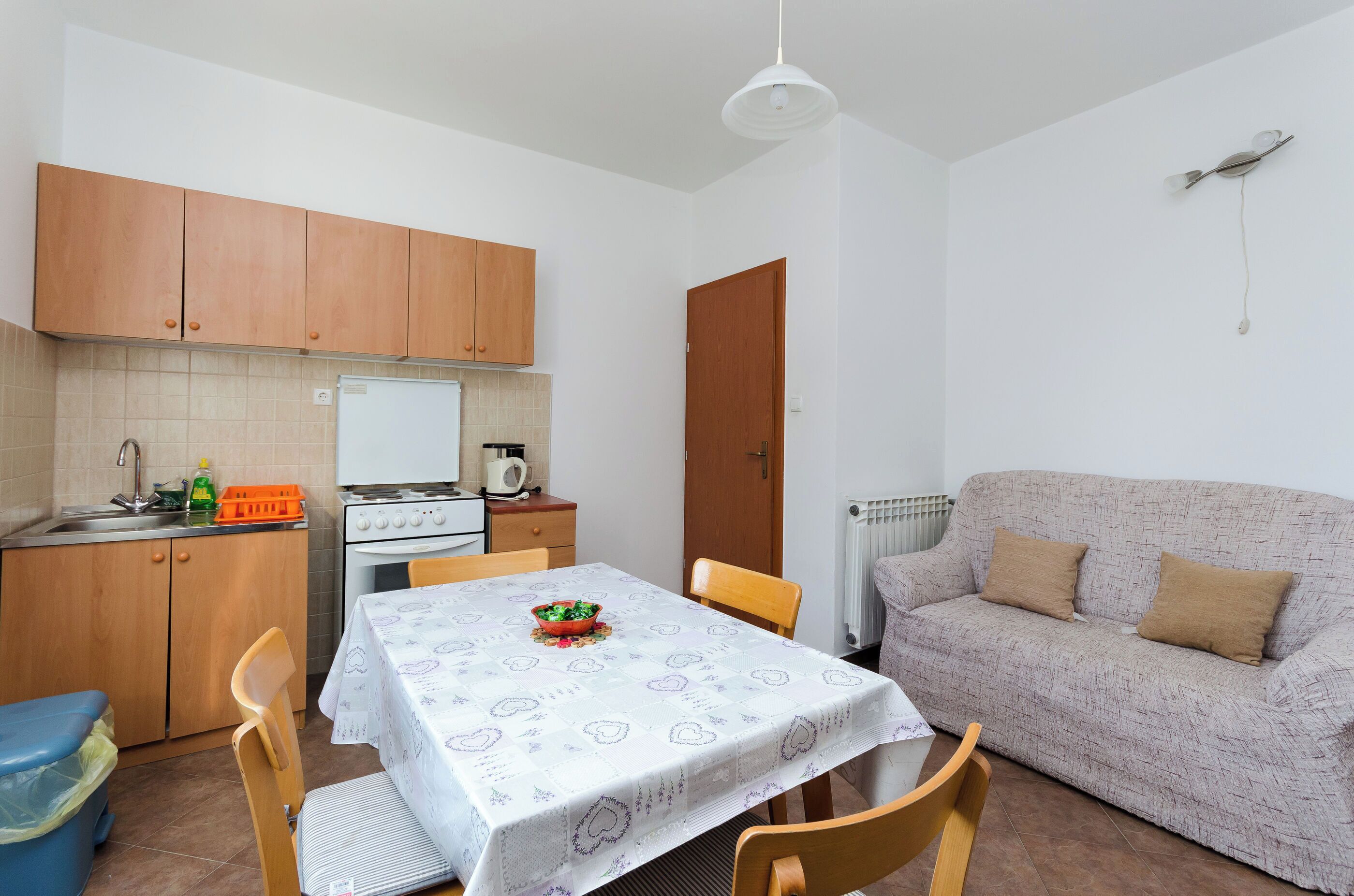 A compact kitchen with wooden cabinets, a dining table with a patterned tablecloth, and a sofa in a living area.