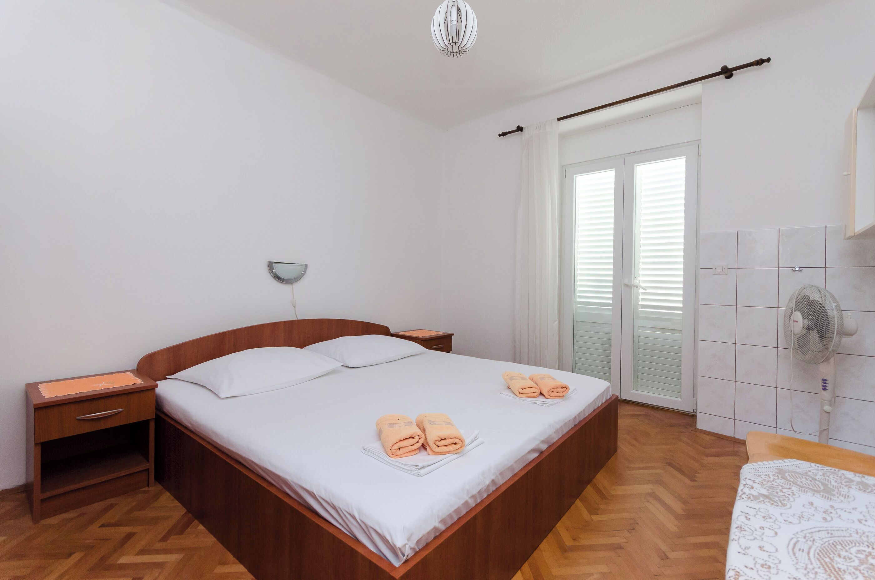 A neatly arranged bedroom with a wooden bed, a bedside table, a nightstand, a wall-mounted fan, and a window with blinds.