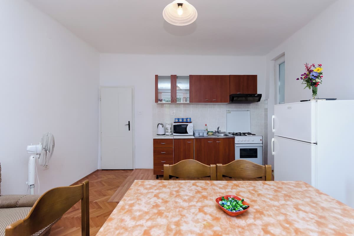 Une cuisine compacte avec des meubles en bois, un four à micro-ondes, une cuisinière et un réfrigérateur. On y trouve une table à manger avec des chaises et un ventilateur dans un coin.