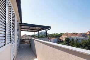 Un balcone con zona salotto riparata, pavimento in pietra e vista su edifici e vegetazione circostanti.