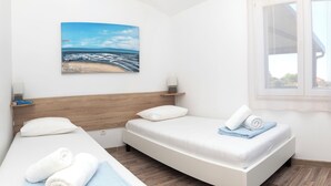 Ein Hotelzimmer mit zwei Betten, einem Holzkopfende, einem Strandbild und einem Fenster mit Aussicht.