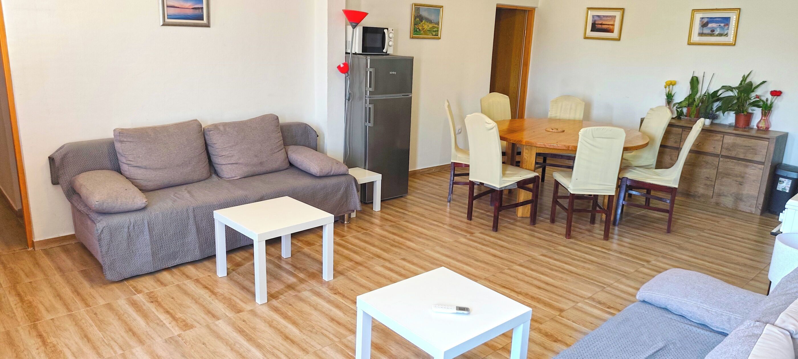 A furnished living room with a sofa, a dining table with chairs, a refrigerator, and a microwave.