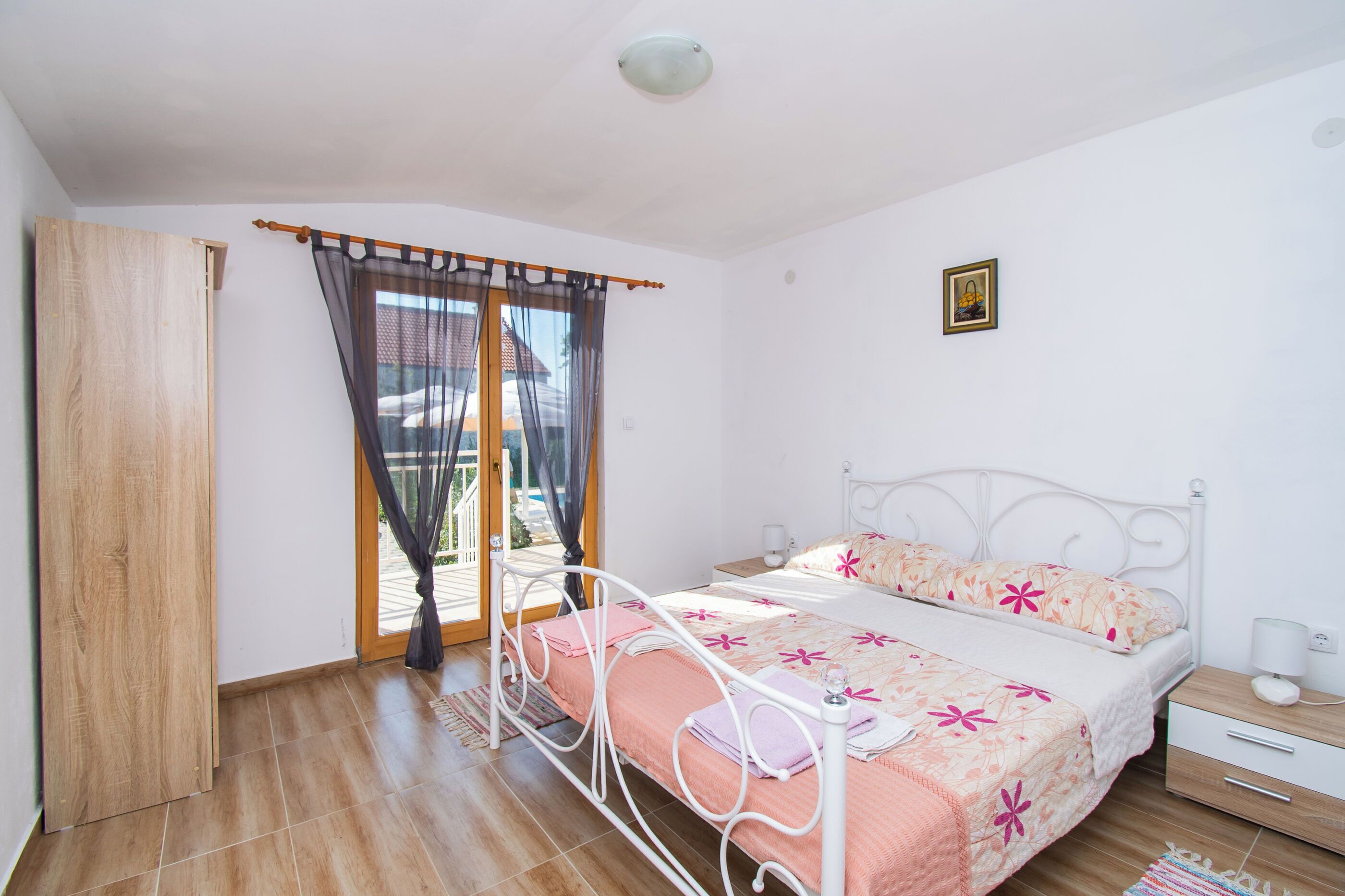 A bedroom with a bed, a wooden door, a window with curtains, a bedside table, and a small framed picture on the wall.