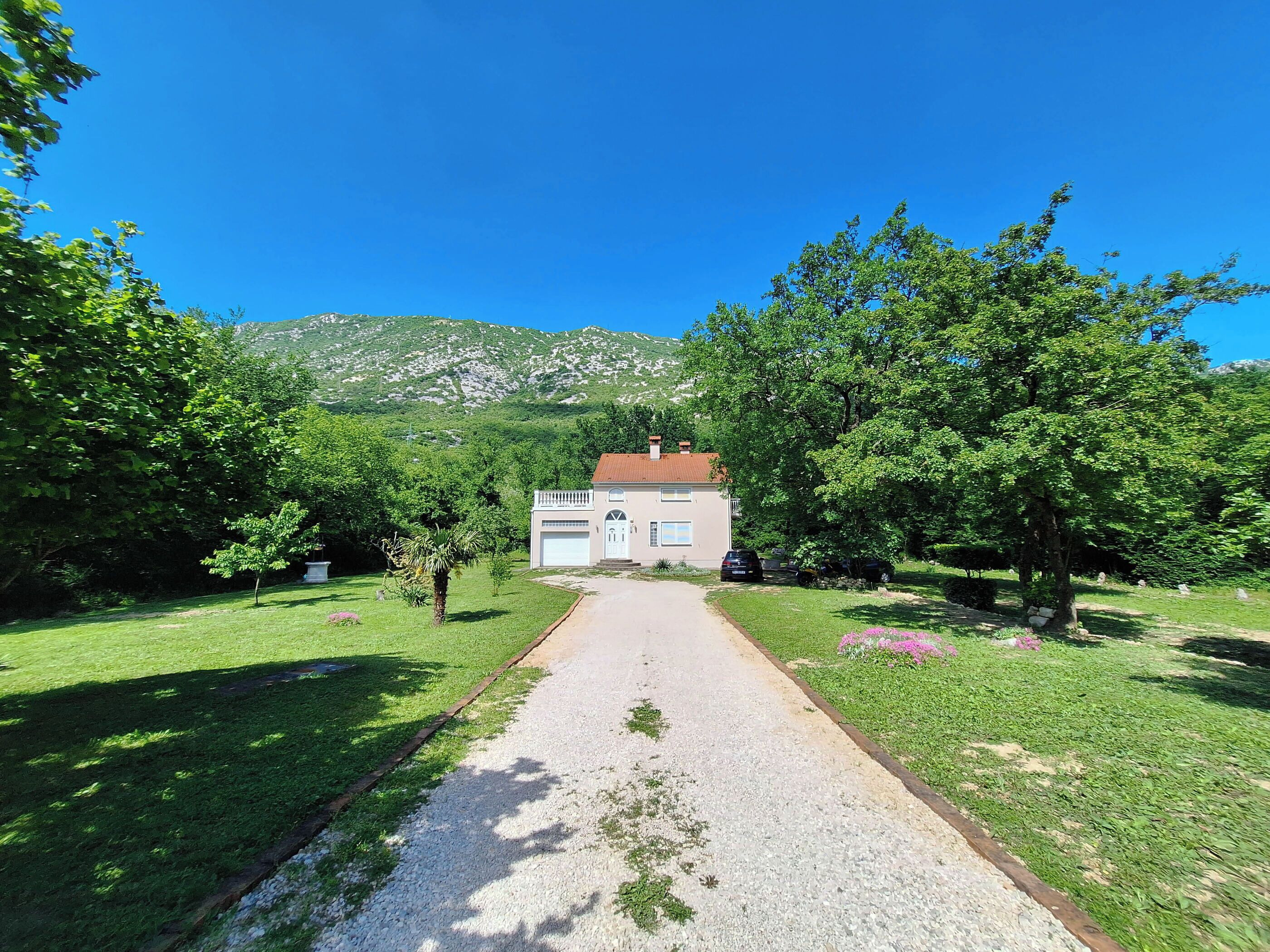 Un sendero conduce a una casa con garaje, rodeada de abundante vegetación y una montaña al fondo.