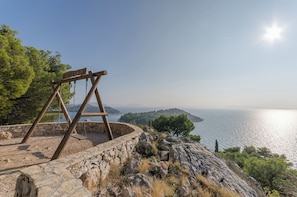 Un columpio de madera sobre una plataforma de piedra con vistas a un paisaje costero con árboles y cielo despejado.