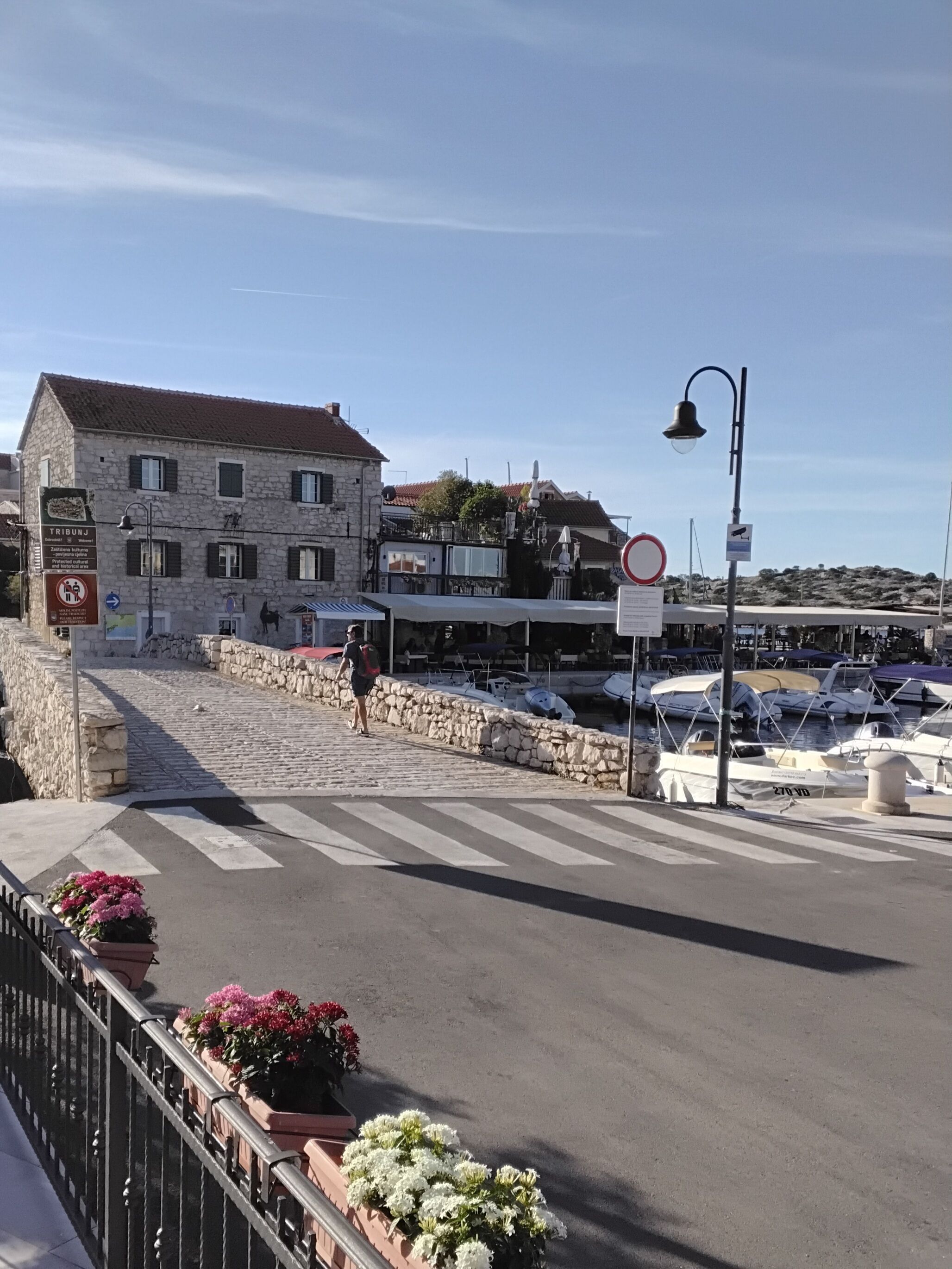A stone building with a signboard, a street with a pedestrian crossing, and a marina with boats.