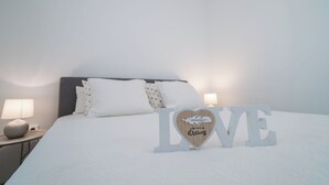 Una cama bien hecha con sábanas y almohadas blancas, una lámpara de noche y letras decorativas que deletrean “LOVE”, con una señal en forma de corazón al frente.