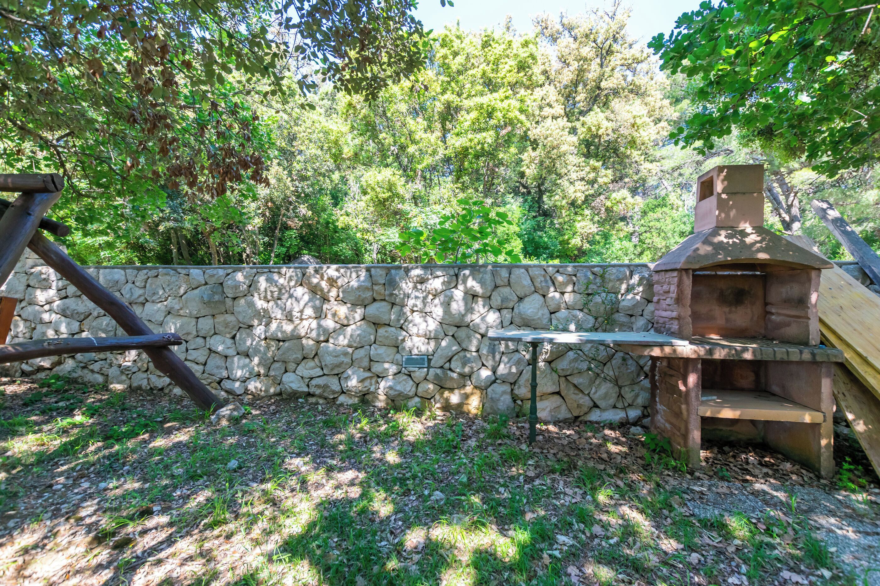 A stone wall, a wooden swing set, and a stone barbecue grill.