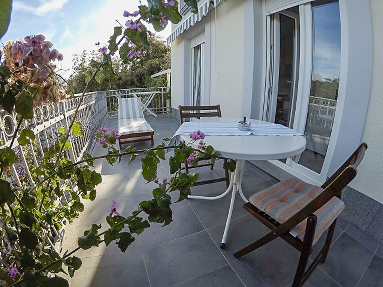 Un balcone con un tavolo rotondo e due sedie, circondato da un parapetto bianco e piante in vaso.