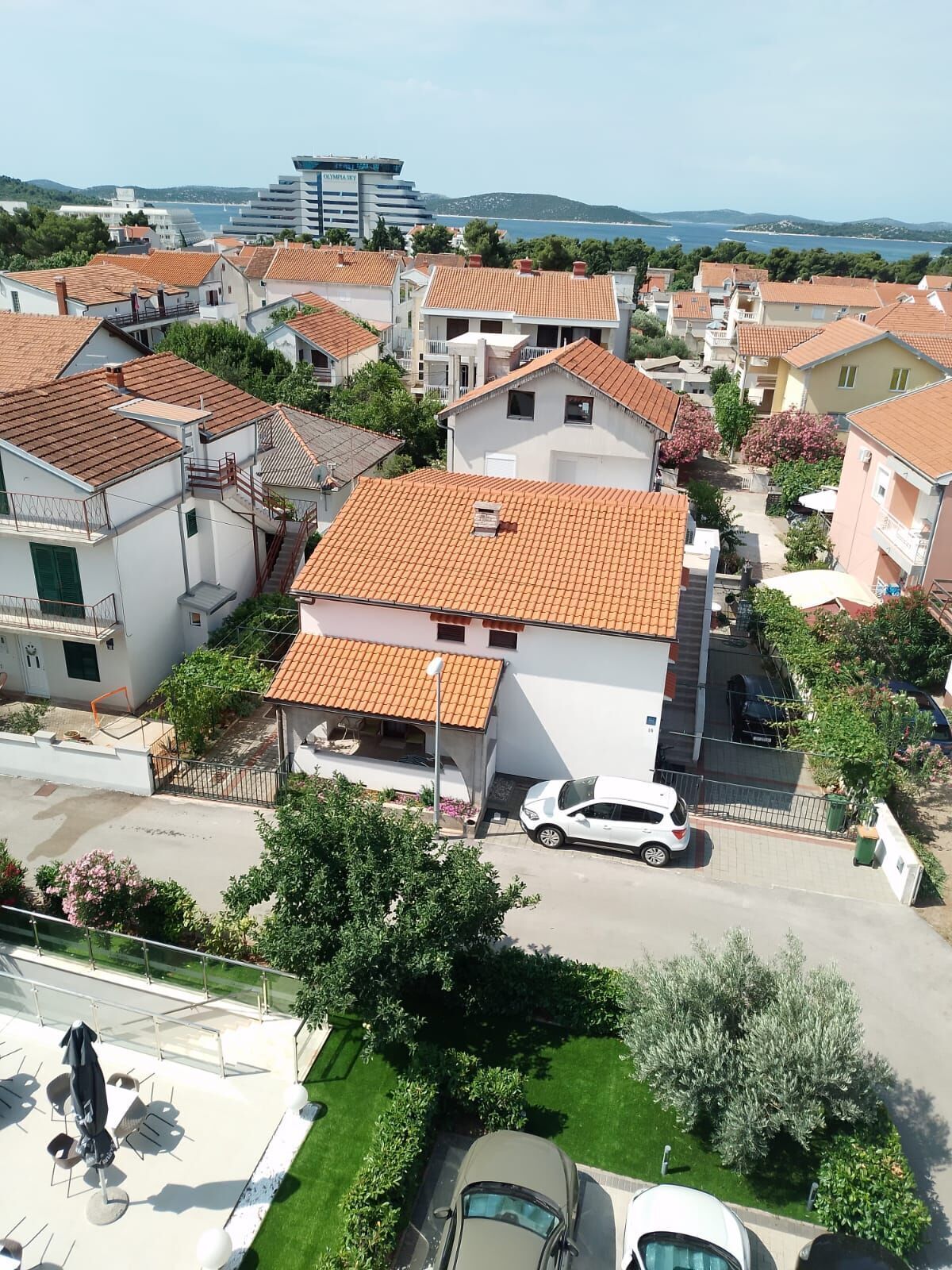 Un quartier résidentiel avec des maisons aux toits recouverts de tuiles rouges, un bâtiment blanc doté de balcons, et une vue sur la mer en arrière-plan.