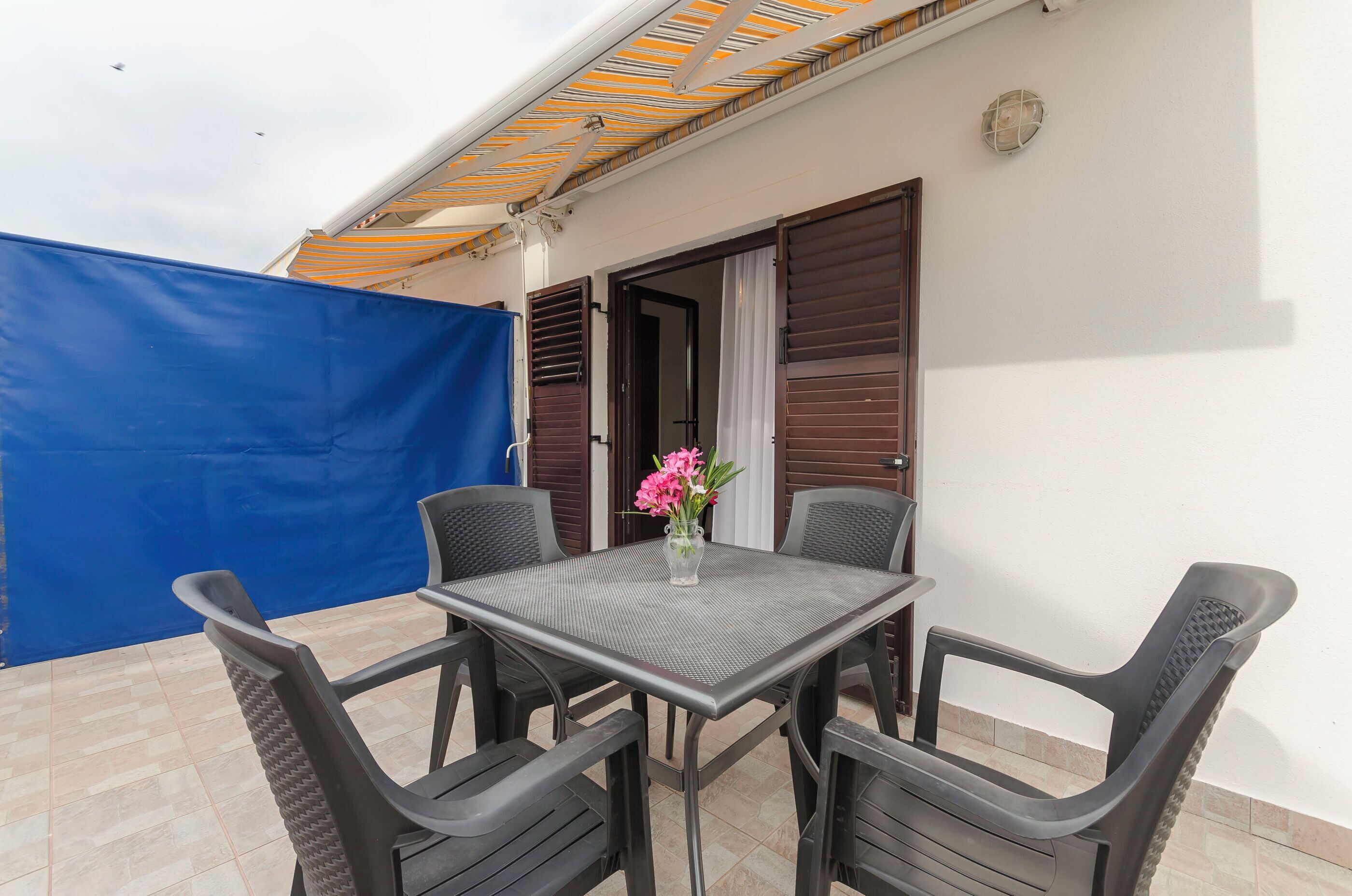 Un patio con una mesa, sillas, un lona azul y una puerta con contraventanas.