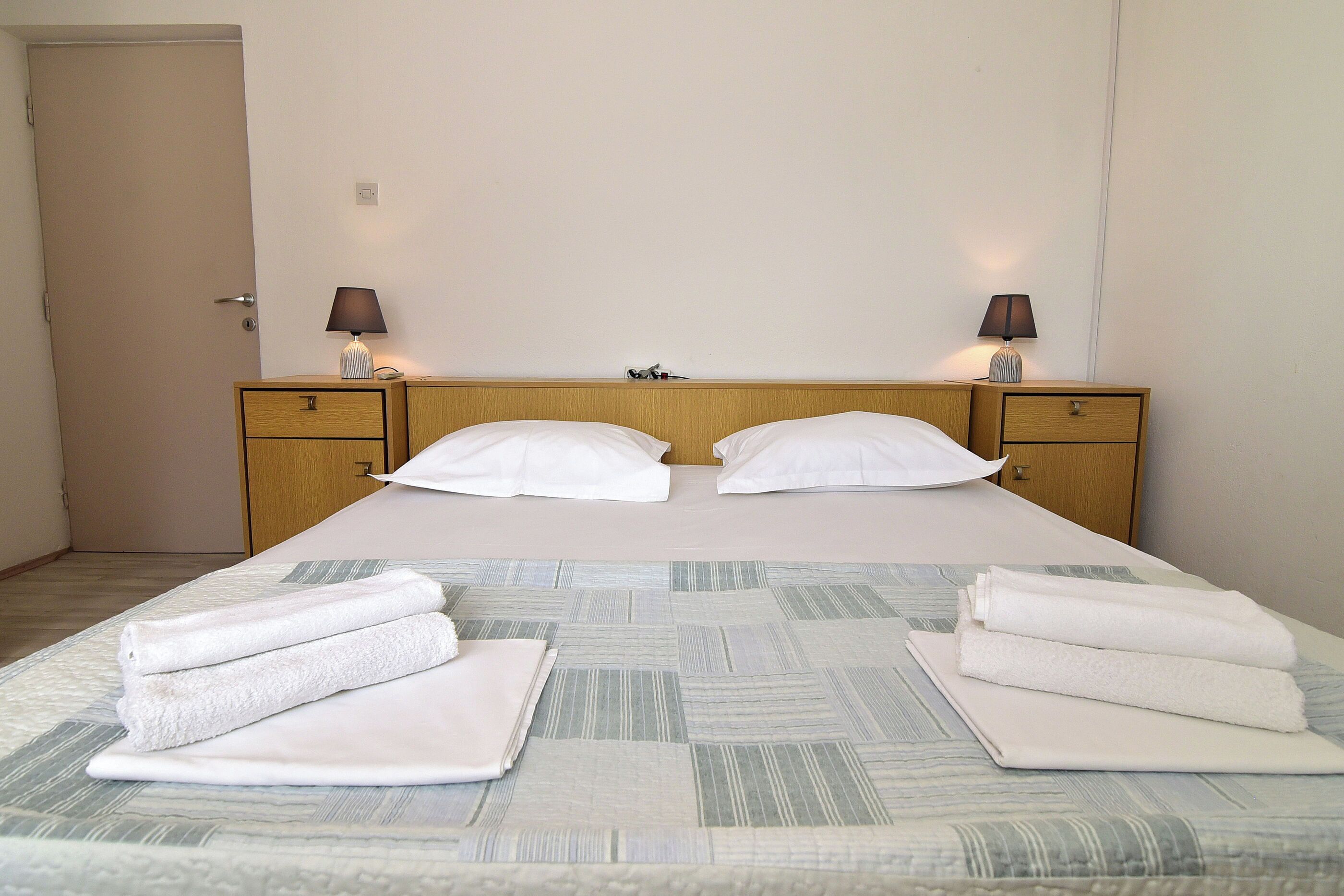 Una cama bien hecha con sábanas blancas, dos mesitas de noche con lámparas y una puerta al fondo.