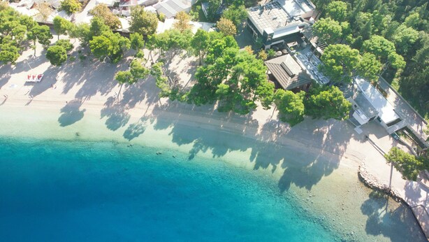 Ett flygbildsbild av ett kustområde med klart blått vatten, en sandstrand och byggnader med sluttande tak.